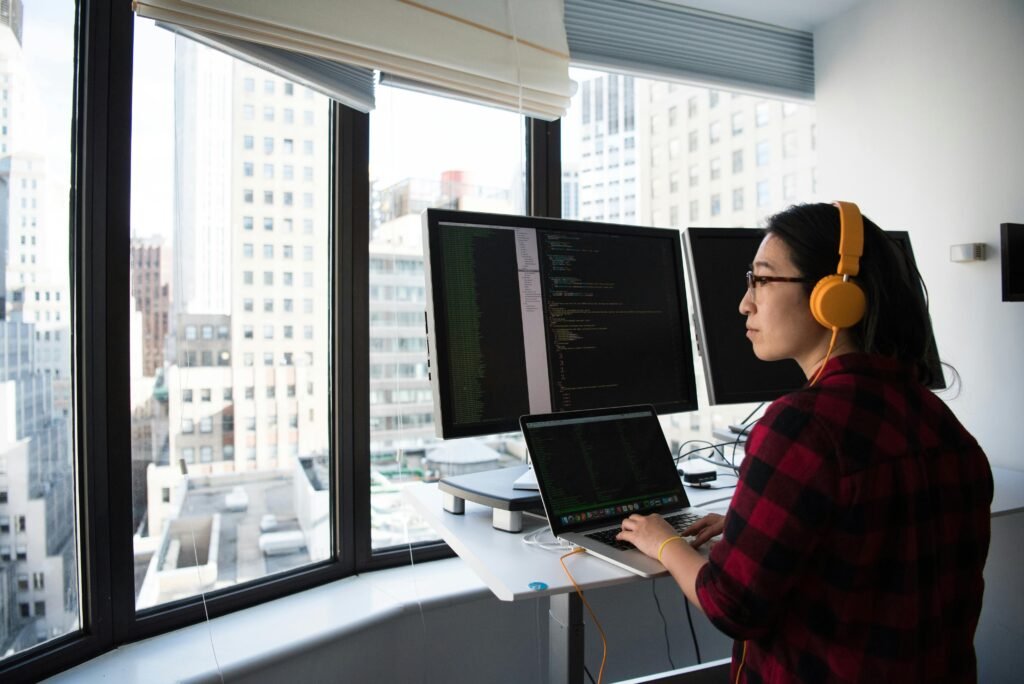 A businesswoman coding at a standing desk with city views through large windows and wearing headphones